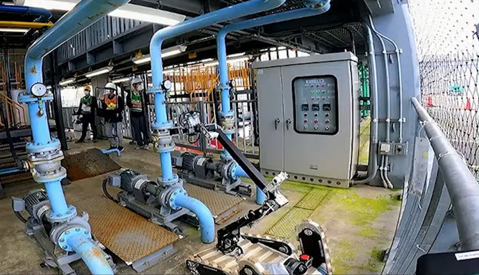 Industrial plant piping and pumps with a tracked inspection robot in the foreground and three helmeted workers observing.